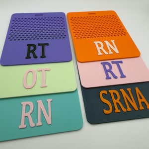 May include: A collection of colorful, rectangular badge holders in shades of purple, orange, pink, teal, green, and navy. Each badge holder has a perforated top and features white or black lettering, including "RT", "RN", "OT", and "SRNA".