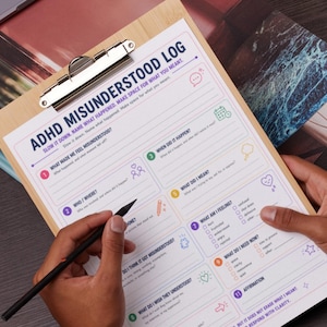 May include: A wooden clipboard holds an ADHD Misunderstood Log. The log is white with colorful sections for recording feelings and thoughts. A black pencil is held over the log, ready to write. The text on the log reads "Slow it down. Name what happened. Make space for what you meant."