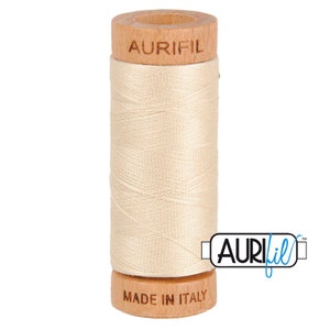 May include: A spool of Aurifil thread in a light beige color. The spool is made of wood and has the Aurifil logo printed on it. The text "Made in Italy" is printed on the bottom of the spool.