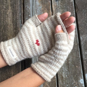 Op de afbeelding: Een paar beige en wit gestreepte vingerloze handschoenen met een rood hartdetail. De handschoenen zijn handgemaakt en hebben een label met de tekst "handgemaakt".