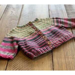 Könnte beinhalten: Ein gestrickter Baby-Cardigan mit Streifen und Knopfleiste. Der Cardigan ist rosa, grün und beige mit einem mehrfarbigen Streifenmuster.