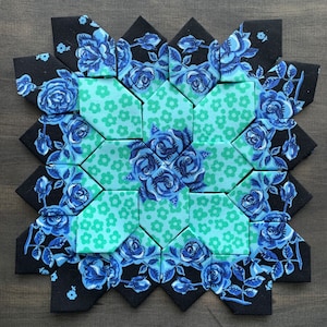 May include: A quilt block with a starburst pattern featuring blue and green fabric. The fabric has a floral design with roses and leaves.