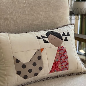 May include: A white pillow with a brown trim features a patchwork design of a stylized person with a red and blue patterned dress and a gray bird with black polka dots.