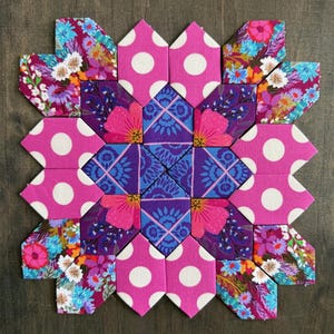 May include: A square fabric quilt block featuring a geometric arrangement of hexagons. The design combines bright pink fabric with white polka dots, purple fabric with blue floral patterns, and multi-colored floral prints in shades of pink, blue, and orange.