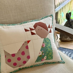 May include: A rectangular throw pillow featuring a pieced quilt design of a person and a chicken. The person has brown hair and a green floral dress. The chicken is beige with pink polka dots. The pillow has a teal floral border trim.