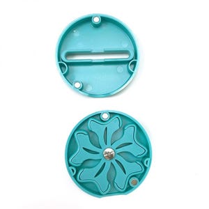 May include: Two-piece teal plastic object. The top piece is a flat circle with a rectangular slot. The bottom piece has a floral design with a central metal piece and magnets. Both pieces have small holes around the edges.