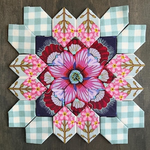 May include: A colorful floral quilt block composed of various fabric patterns. The center features a large, stylized flower in shades of pink, purple, and red. Surrounding the flower are light blue floral and gingham patterned pieces.