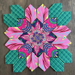 May include: A colorful, floral-patterned fabric coaster. The coaster features a central flower design in pink, blue, and purple, surrounded by green and pink geometric shapes. The design is set against a dark wood background.