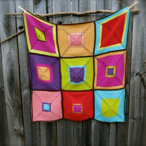 Pode incluir: Uma manta de malha quadrada e colorida com um design geométrico. A manta apresenta nove quadrados em várias cores, incluindo rosa, vermelho, laranja, roxo e verde, cada um com um quadrado central contrastante e bordas marrons escuras. É exibida contra uma cerca de madeira.
