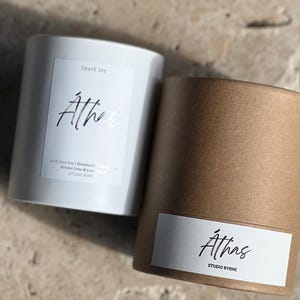 May include: Two cylindrical candles, one white and one brown, with labels. The white candle reads "Spark Joy" and "Athas." The brown candle reads "Athas STUDIO BYRNE." The white candle also lists ingredients and "STUDIO BYRNE."
