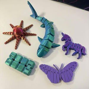 Puede incluir: Una colección de juguetes articulados impresos en 3D, coloridos. Incluye un tiburón, un pulpo, una mariposa, un unicornio y un bloque segmentado. El tiburón es verde azulado y azul, el pulpo es rojo y dorado, y la mariposa es morada.