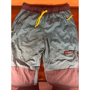 May include: Teal and purple outdoor pants with a drawstring waist and yellow accents. The pants have a Berghaus logo and pockets. The lower portion of the pants is a darker purple color.