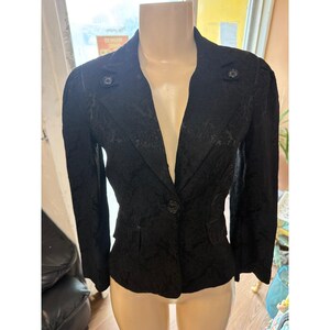 Peut inclure: Veste noire avec un motif texturé, présentée sur un mannequin. La veste a un col cranté, deux poches avant et des boutons décoratifs. Elle a des manches longues et une fermeture à un seul bouton.
