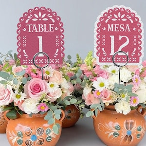 May include: Two terracotta pots filled with pink and white floral arrangements, each featuring a scalloped-edge cardstock sign. One sign displays the text TABLE 1, and the other displays MESA 12, both printed in white on a deep red background.