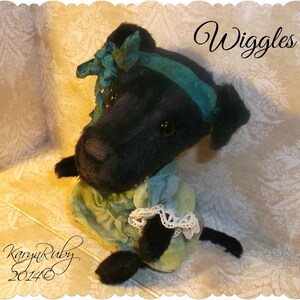 Könnte beinhalten: Schwarzes Plüschtierhund, das ein blaugrünes Kleid mit weißer Spitzenborte und einer blaugrünen Schleife trägt. Der Hund sitzt auf einem weißen und goldfarbenen gemusterten Stoff. Der Text "Wiggles" ist in Schwarz über dem Hund geschrieben.