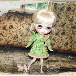Könnte beinhalten: Eine Blythe-Puppe mit blondem Haar und blauen Augen, die ein grün kariertes Kleid mit Rüschenkragen trägt. Die Puppe steht in einem Raum mit floraler Tapete und einer kleinen Puppe auf dem Boden. Das Gesicht der Puppe ist mit rosigen Wangen bemalt.