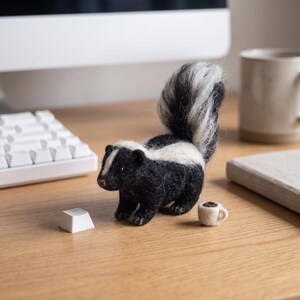 May include: A needle-felted skunk figurine with black and white wool fibres, featuring a prominent upright tail, a white stripe along its back and head, and small dark eyes. Next to the skunk is a miniature beige mug and a single white keycap.