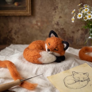 Könnte beinhalten: Eine nadelgefilzte Fuchsfigur aus orangefarbenen, weißen und schwarzen Wollfasern, die in einer Schlafposition zusammengerollt ist. Der Fuchs hat spitze Ohren mit schwarzen Spitzen, eine weiße Brust und einen weiß gefärbten Schwanz. Nadel und Skizze.