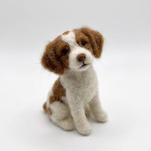 May include: A handcrafted felt dog figurine in a sitting position. The dog has brown and white fur, with brown ears and patches on its body. The dog has a small, brown nose and is set against a white background.