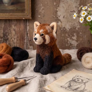 May include: A needle-felted red panda figurine with a textured brown and black coat, cream-coloured facial markings, and pointed ears. The small animal is crafted from wool fibres and sits upright, featuring dark eyes and a black nose.