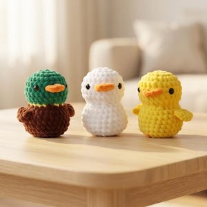 Könnte beinhalten: Drei gehäkelte Enten in einer Reihe. Eine ist grün und braun, eine weiß und die andere gelb. Jede hat einen orangefarbenen Schnabel und kleine schwarze Augen. Die Enten sitzen auf einem hellen Holztisch.