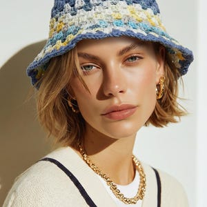 Op de afbeelding: Een gehaakte bucket hat in blauw, geel en wit. De hoed heeft een brede rand en een textuur. Het model draagt een crèmekleurige trui met marineblauwe bies en een gouden ketting.