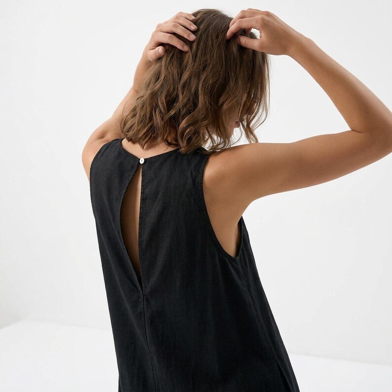 May include: Black sleeveless dress with a keyhole back and a single button closure at the neckline. The dress is made of a textured fabric and has a simple, elegant design. The model's hands are in their hair.