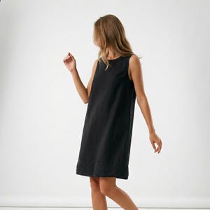 May include: A black sleeveless dress with a minimalist design. The dress features a round neckline and falls to the mid-thigh. The fabric looks to be a lightweight material, ideal for warmer weather. The dress is shown on a person.