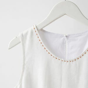 May include: White sleeveless top with a round neckline and brown stitched detailing. The top is displayed on a white wooden hanger. The fabric looks like linen or a similar lightweight material.