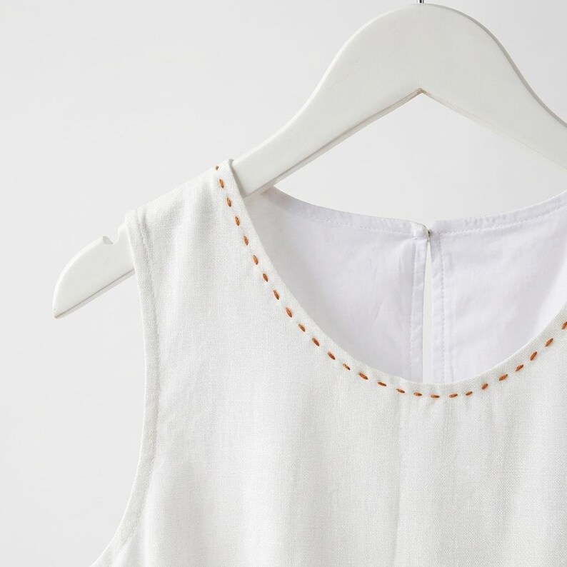 May include: White sleeveless top with a round neckline and brown stitched detailing. The top is displayed on a white wooden hanger. The fabric looks like linen or a similar lightweight material.