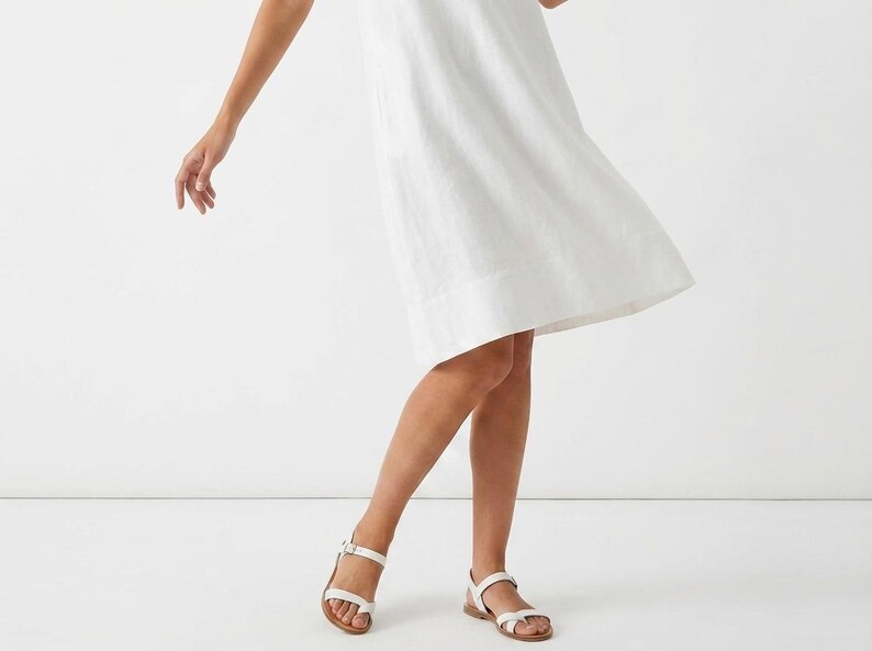 May include: A white linen dress with a simple design. The dress is paired with white leather sandals with a buckle closure. The sandals have a brown sole. The image is set against a white background.