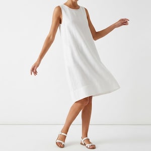 May include: A white linen dress with a round neckline and no sleeves. The dress reaches the mid-calf and is worn with white sandals. The model is captured in motion, suggesting a breezy, casual style.