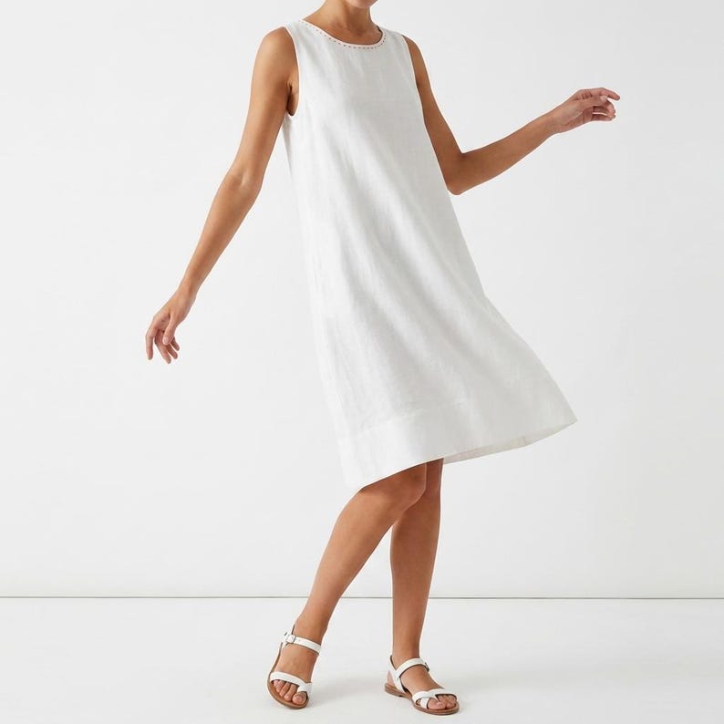 May include: A white linen dress with a round neckline and no sleeves. The dress reaches the mid-calf and is worn with white sandals. The model is captured in motion, suggesting a breezy, casual style.