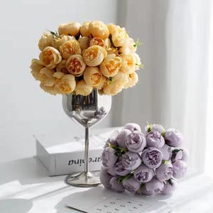 May include: A still life image showing two bunches of artificial flowers. One is a bouquet of yellow flowers in a silver goblet vase. The other is a cluster of lavender-coloured flowers.