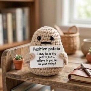 May include: A crocheted potato figure with a neutral color scheme, holding a sign that reads "Positive potato." The sign continues with the text "I may be a tiny potato, but I believe in you. Go do your thing!" The figure is on a wooden shelf.
