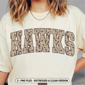 May include: A cream-colored short-sleeve t-shirt featuring the word HAWKS in a large, arched collegiate font. The letters have a brown, tan, and green camouflage pattern. A small rectangular label at the bottom reads 2 - PNG FILES - DISTRESSED & CLEAN VERSION.