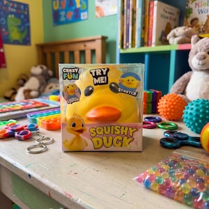 May include: A yellow squishy duck toy in a clear plastic package with the text "Crazy Fun" and "Try Me!". The box also features a cartoon duck illustration. Other toys are scattered on a white table.