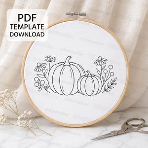 Può includere: Un telaio da ricamo rotondo in legno contenente tessuto bianco con un motivo a linee nere di due zucche circondate da fiori di campo e steli. Un riquadro bianco nell'angolo in alto a sinistra contiene il testo PDF TEMPLATE DOWNLOAD.