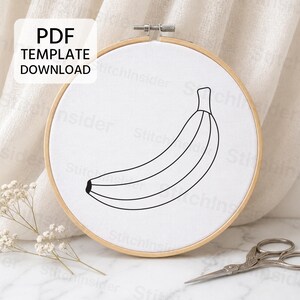 May include: A circular wooden embroidery hoop containing white fabric with a black line art drawing of a banana. A white box in the upper left corner contains the black text PDF TEMPLATE DOWNLOAD. The hoop is positioned for display.