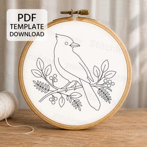 May include: A wooden embroidery hoop containing white fabric with a black line art pattern of a bird perched on a leafy branch with berries. A white spool of thread sits to the left. A white box with the text PDF TEMPLATE DOWNLOAD is visible.