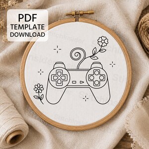 May include: A circular wooden embroidery hoop containing white fabric with a black line art design of a video game controller. A flower grows from the top of the controller, and another flower sits to the left. The text PDF TEMPLATE DOWNLOAD is visible.
