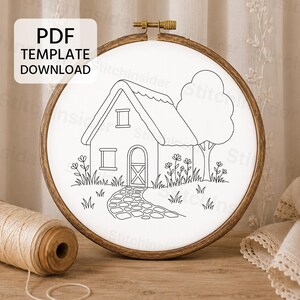 May include: A wooden embroidery hoop containing white fabric with a black line art pattern of a cottage, a tree, flowers, and a stone path. A text box in the upper left corner reads PDF TEMPLATE DOWNLOAD. A spool of beige thread sits nearby.