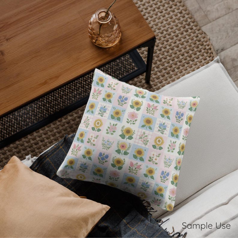 Op de afbeelding: Vierkant sierkussen met een bloemenpatroon met zonnebloemen, blauwe bloemen en roze bloemen op een lichtroze en blauwe geruite achtergrond. Het kussen ligt op een witte bank met een houten tafel op de achtergrond.