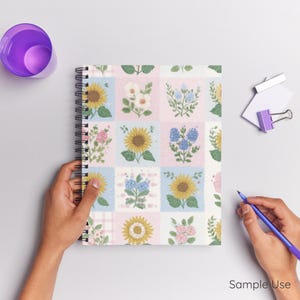 Op de afbeelding: Een spiraalgebonden notitieboek met een bloemenpatchworkontwerp met zonnebloemen en andere bloemen in pastelkleurige vierkanten. De woorden "Sample Use" staan rechtsonder afgedrukt.