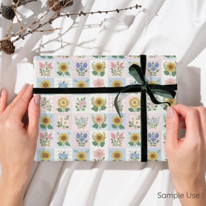 Op de afbeelding: Een cadeau verpakt in bloemenpatroonpapier met zonnebloemen en andere bloemen in een rasterontwerp. Het is vastgebonden met een zwart fluwelen lint en een groene strik. De woorden "Sample Use" zijn zichtbaar.