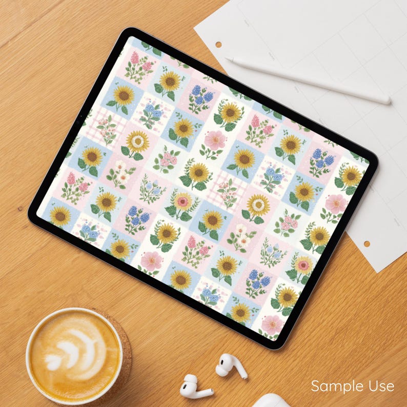 Op de afbeelding: Een digitale tablet toont een bloemenpatroon met zonnebloemen, roze rozen en blauwe bloemen op een witte en pastelkleurige achtergrond. De tablet ligt op een houten oppervlak met een kopje koffie, oortelefoons en een potlood.