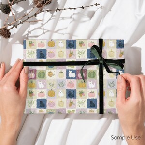 May include: A rectangular gift wrapped in patterned paper featuring a grid of illustrations including black and white cats, pumpkins in shades of yellow, green, and pink, and sprigs of greenery, tied with a dark green velvet ribbon.