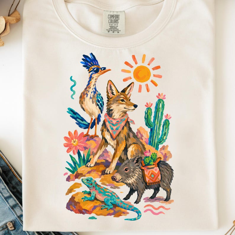 May include: A cream-colored t-shirt featuring a colorful graphic print of a roadrunner wearing sunglasses, a coyote with a patterned bandana, a javelina with a saddlebag, a lizard, a cactus, a bright sun, and desert flowers.
