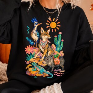 May include: A black crewneck sweatshirt featuring a colorful graphic print of a desert scene. The design includes a roadrunner, a coyote wearing a patterned bandana, a javelina with a saddlebag, a lizard, a cactus, and a bright yellow sun.