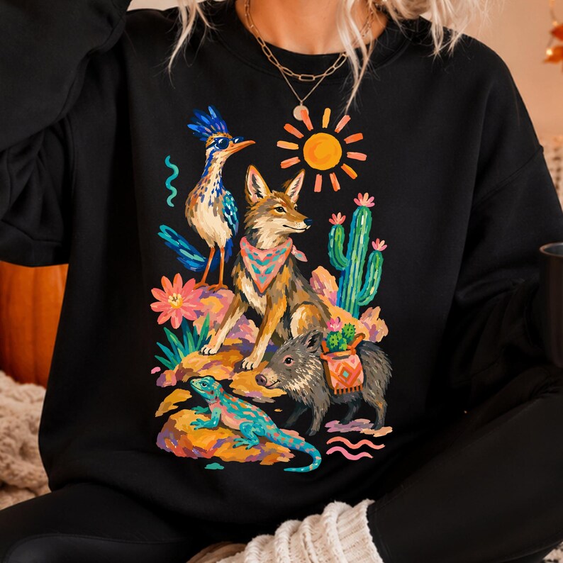 May include: A black crewneck sweatshirt featuring a colorful graphic print of a desert scene. The design includes a roadrunner, a coyote wearing a patterned bandana, a javelina with a saddlebag, a lizard, a cactus, and a bright yellow sun.